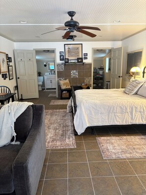 A bedroom with a bed, a chair, a ceiling fan, and a bathroom visible in the background.