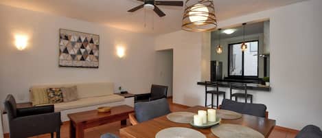 A modern living room with a dining area, a ceiling fan, and a kitchen visible through an open doorway.