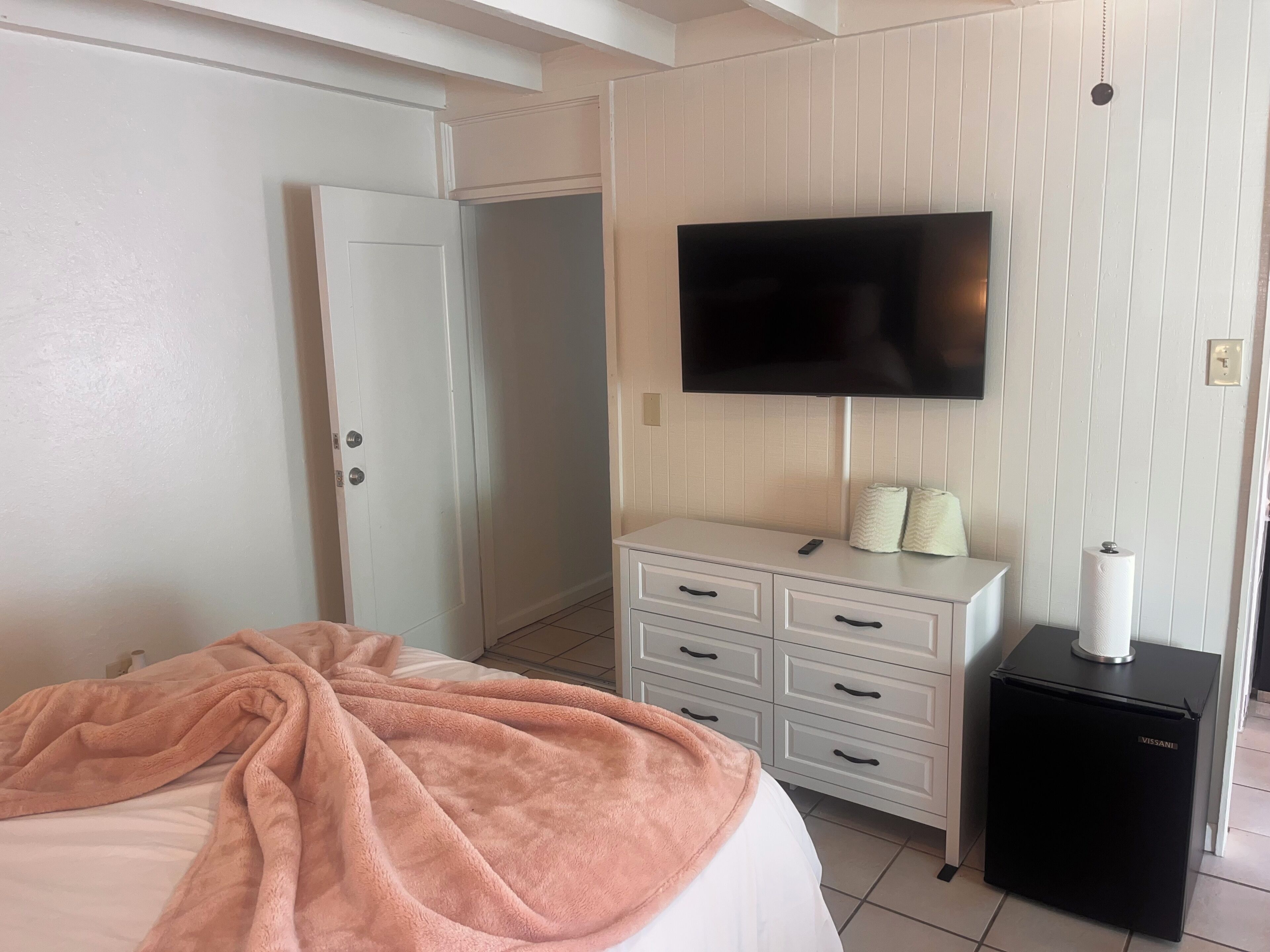 A hotel room with a bed, a television mounted on the wall, a white dresser with drawers, and a small black refrigerator.
