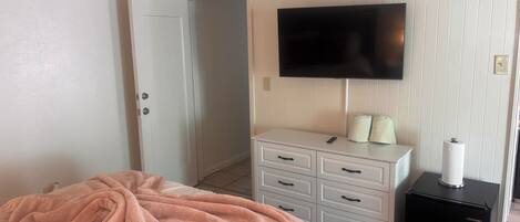 A hotel room with a bed, a television mounted on the wall, a white dresser with drawers, and a small black refrigerator.