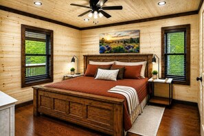 A wooden-paneled bedroom with a large bed, two bedside tables, a ceiling fan, and a painting above the bed. - Sweet Sage Lodge -New Pool December 2026! (Broken Bow)