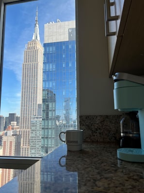 A kitchen with a coffee maker and a mug on the counter, a window with a view of a tall building, and a granite countertop.