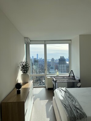 A bedroom with a large window, a bed, a bedside table, a dresser, and a potted plant.