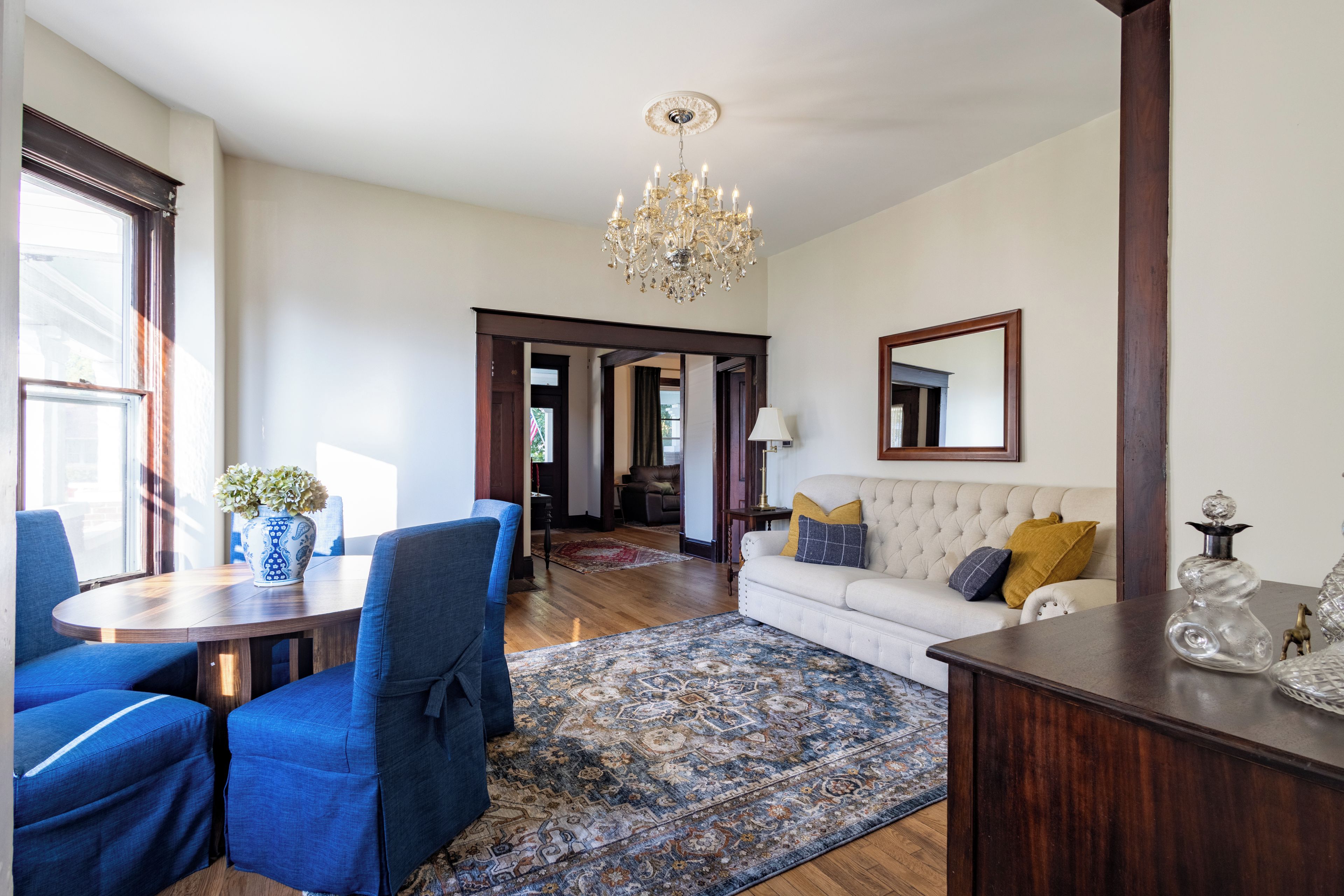 Un salon spacieux agrémenté d’un lustre, un coin repas avec des chaises bleues, un canapé orné de coussins décoratifs et un grand tapis.