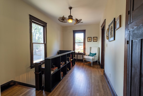 A hallway with wooden flooring, a black bookshelf, a chair with a green cushion, and a ceiling fan.