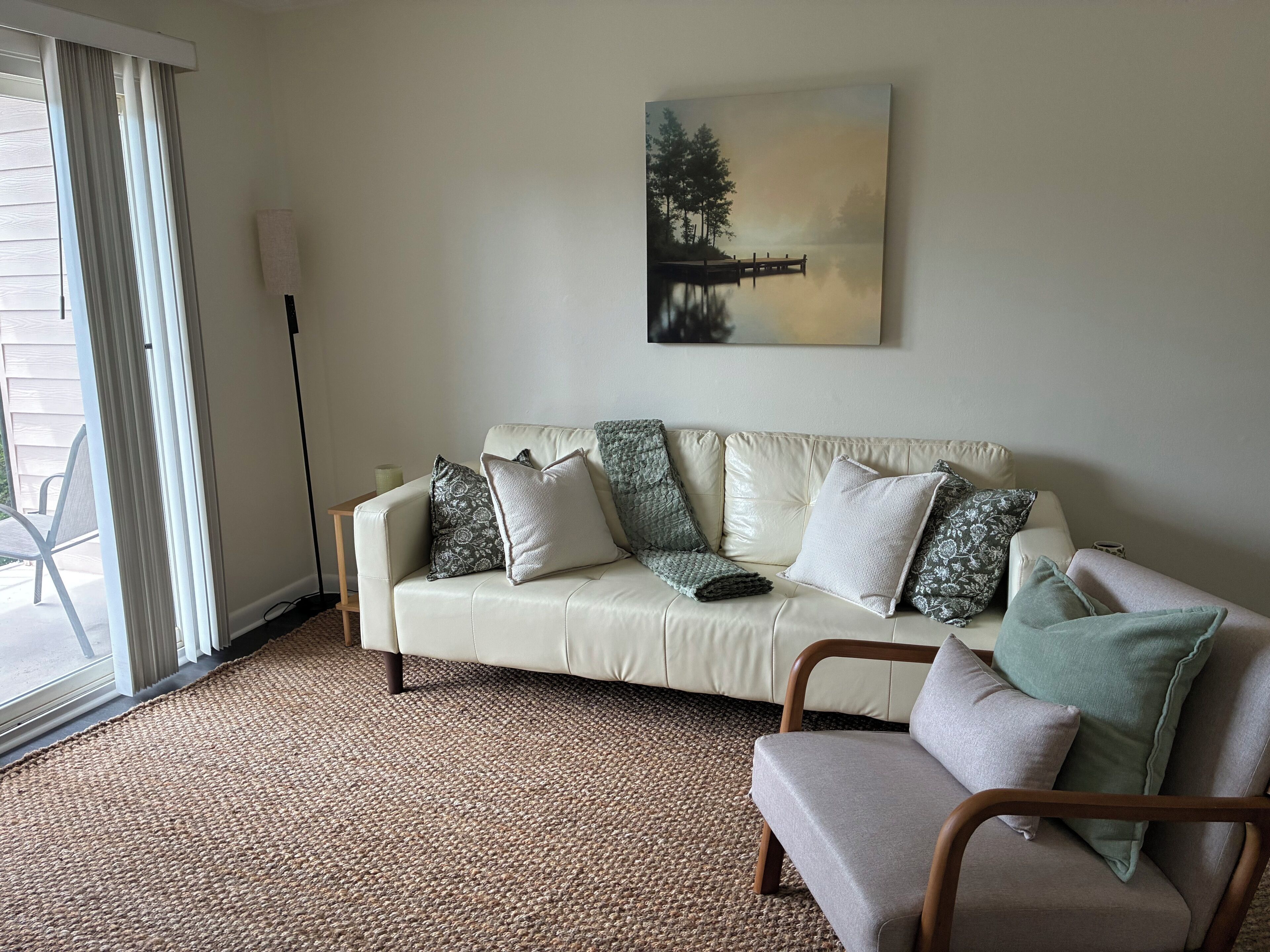 A living room with a beige sofa, a green armchair, and a painting of a lake scene.
