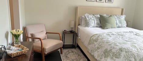 Una habitación de hotel con cama, una silla, una mesita de noche, una lámpara, un jarrón con flores y cuadros en la pared.