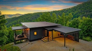 Ein modernes Haus mit einer großen Terrasse und einem überdachten Außenbereich, umgeben von bewaldeten Hügeln.
