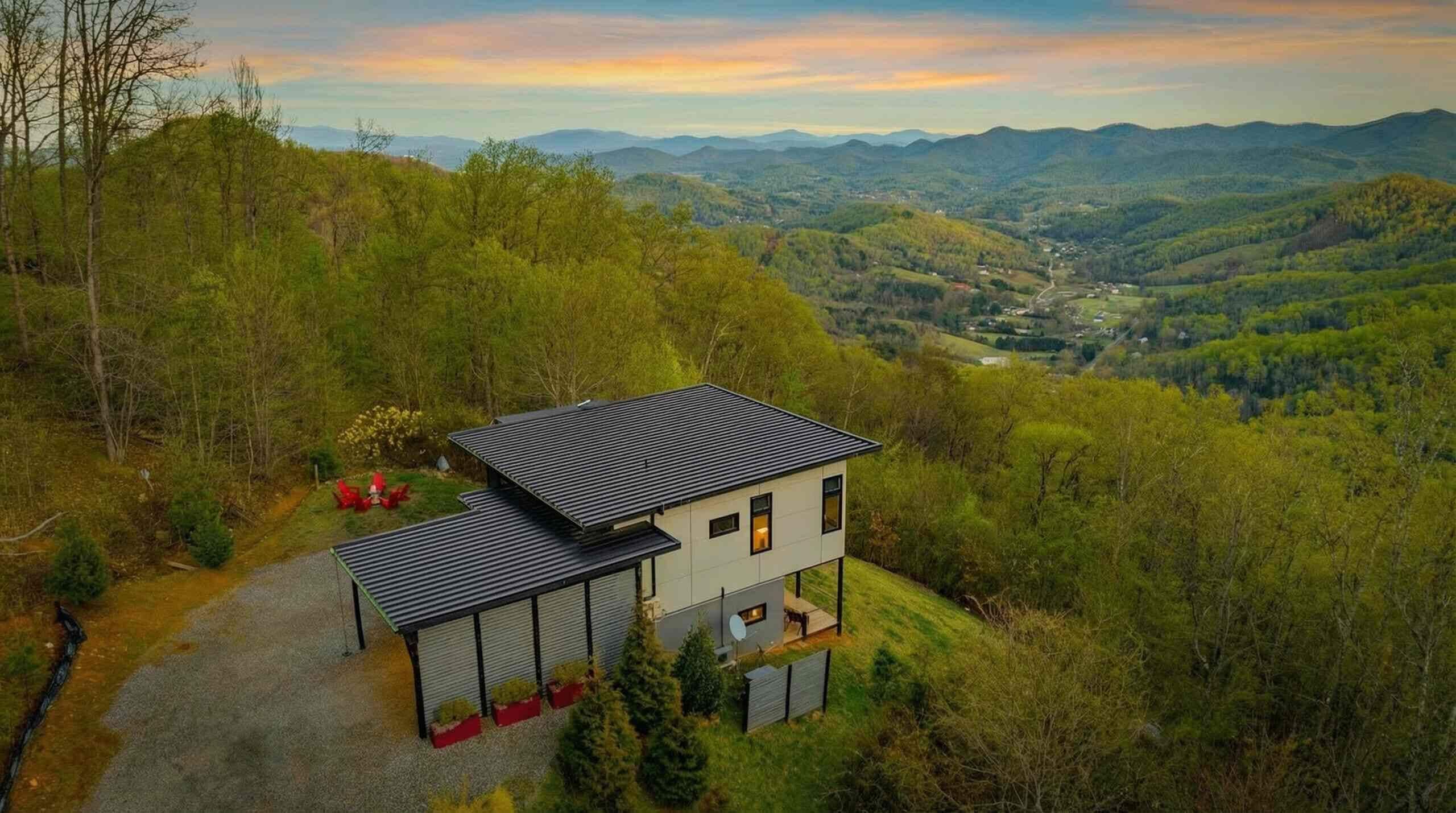 A modern two-story house with a flat roof, surrounded by lush greenery and hills in the background.