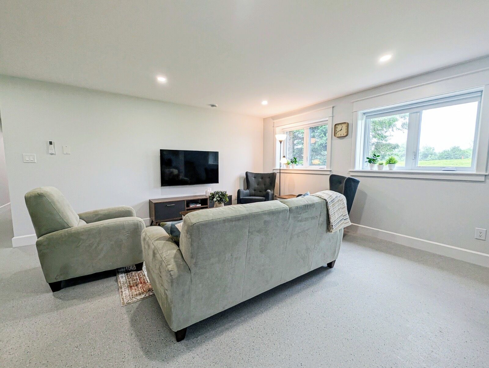 A living room with a grey sofa, two armchairs, a flat-screen TV, a small table with a lamp, and a window with a view of greenery.