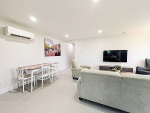 A modern living room with a sofa, a dining table with chairs, a flat-screen TV, and an air conditioning unit on the wall.