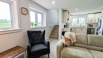 Un espace de vie compact comprenant un canapé, un fauteuil et une petite table. On aperçoit également un coin cuisine avec des placards et des appareils électroménagers.