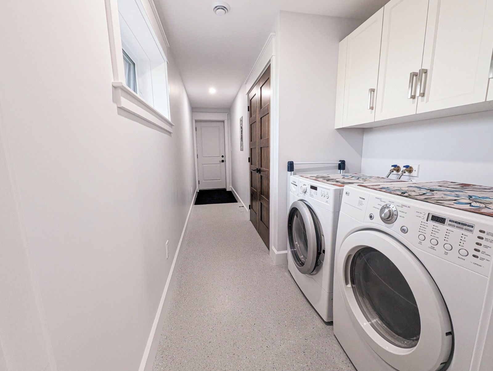 A laundry room with two front-loading washing machines and a dryer, a door leading to another room, and a window on the left wall.