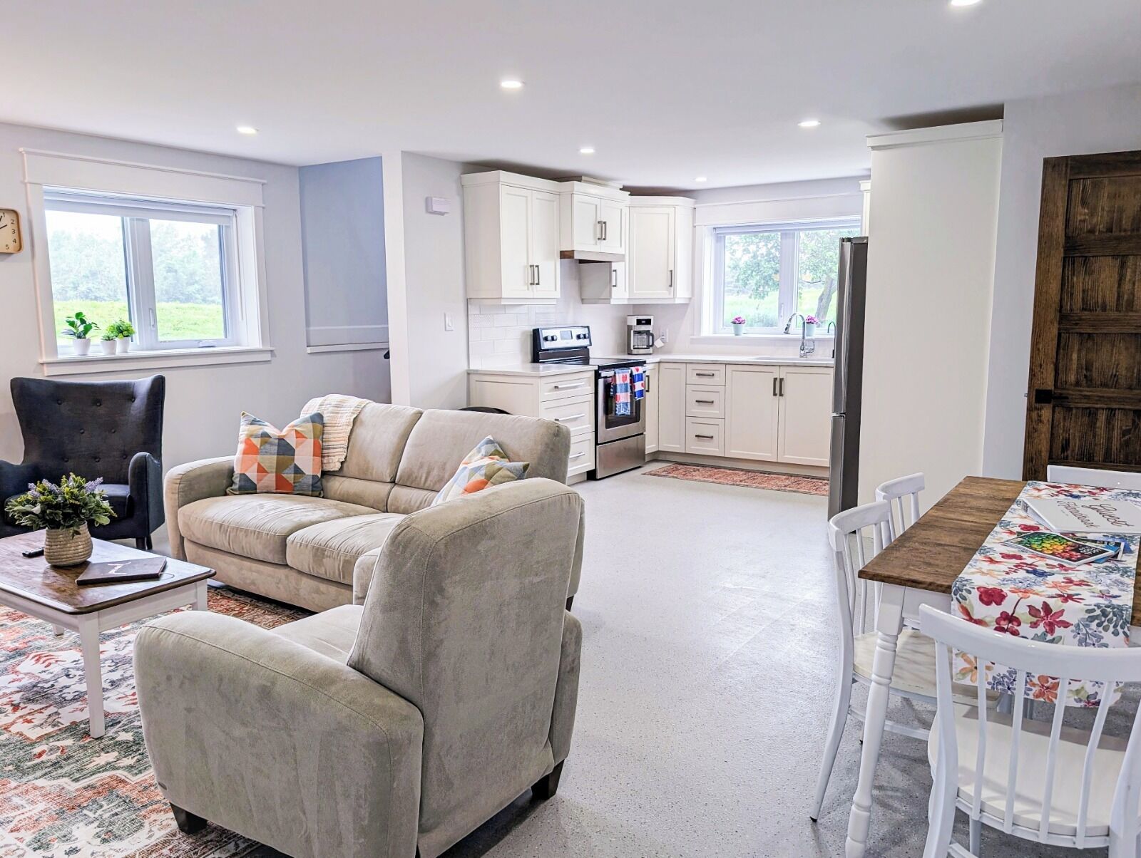 A modern living room with a sofa, a dining table with chairs, a kitchen area with cabinets and appliances, and a view of the outdoors through the windows.