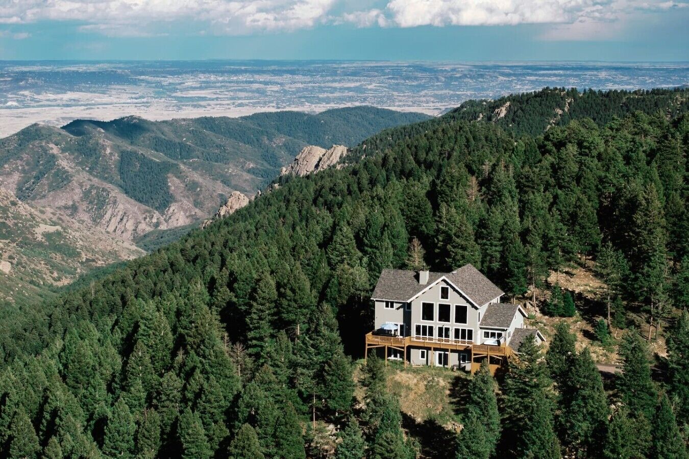 A large house with a deck, surrounded by dense forest and mountains.