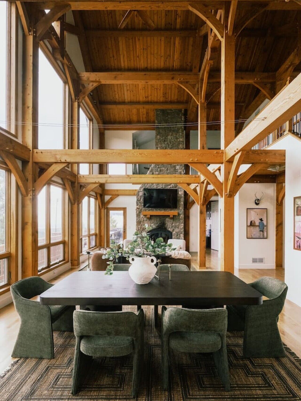 Un'ampia sala da pranzo con soffitto alto e travi a vista in legno, un camino in pietra e vista all'esterno attraverso ampie finestre.