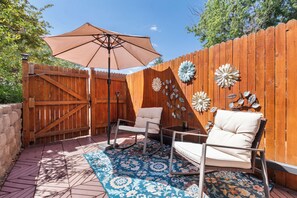 Eine Terrasse mit Holzzaun, Klapptisch, zwei Stühlen, einem gemusterten Teppich und dekorativen Metallblumen.