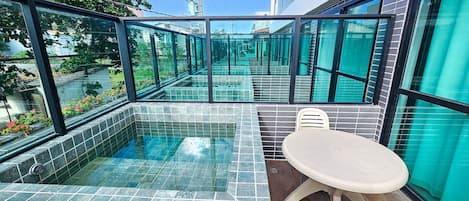 Un área de piscina moderna al aire libre con paredes de vidrio, piso de baldosas y una mesa con sillas.