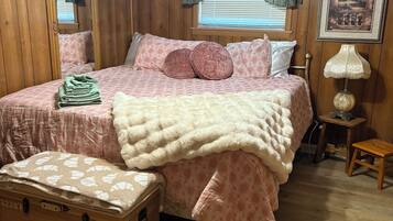 Un dormitorio con piso de madera, una cama con ropa de cama rosa, un espejo, una lámpara y una maleta.