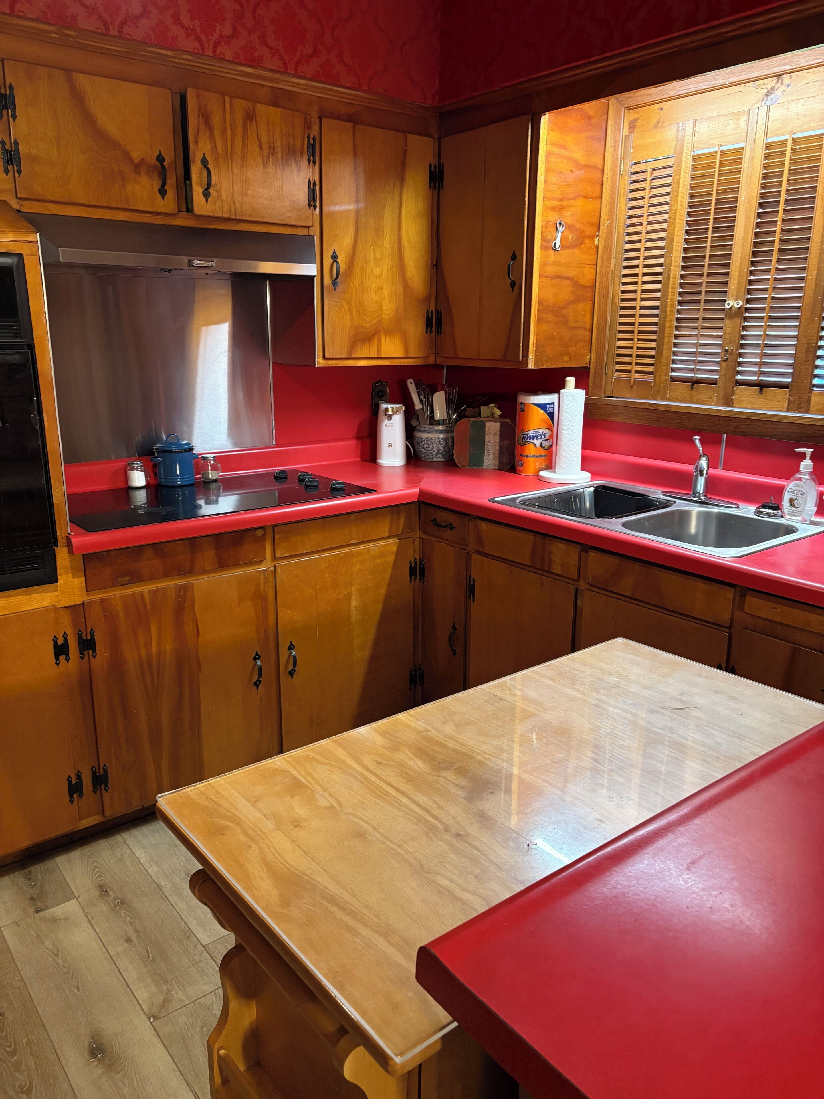 Una cocina con armarios de madera, encimera roja, pileta de acero inoxidable y ventana con contraventanas.