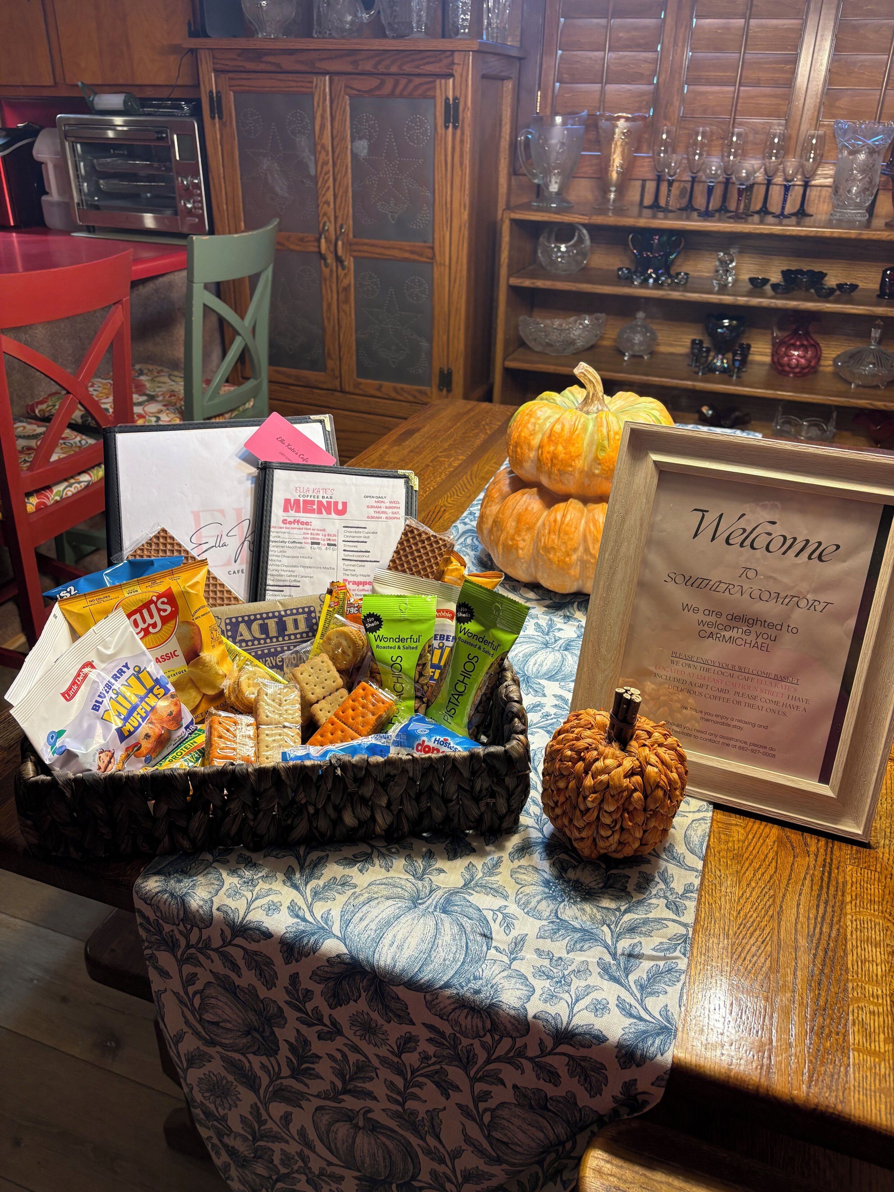 Un cartel de bienvenida, una cesta de aperitivos y una calabaza sobre una mesa en una habitación acogedora con muebles de madera y un mantel estampado.