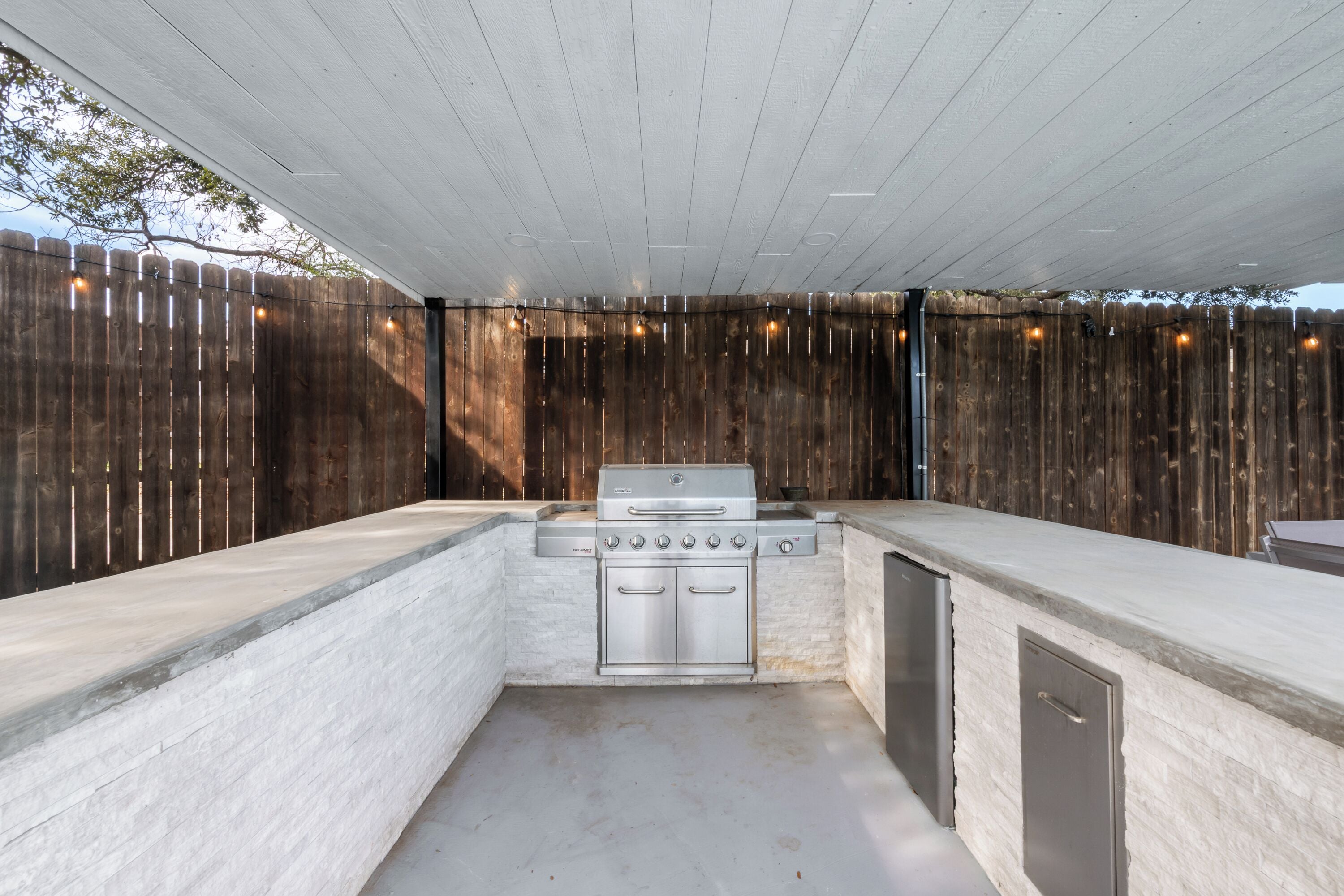An outdoor kitchen with a built-in grill and a dishwasher, surrounded by a wooden fence and string lights.