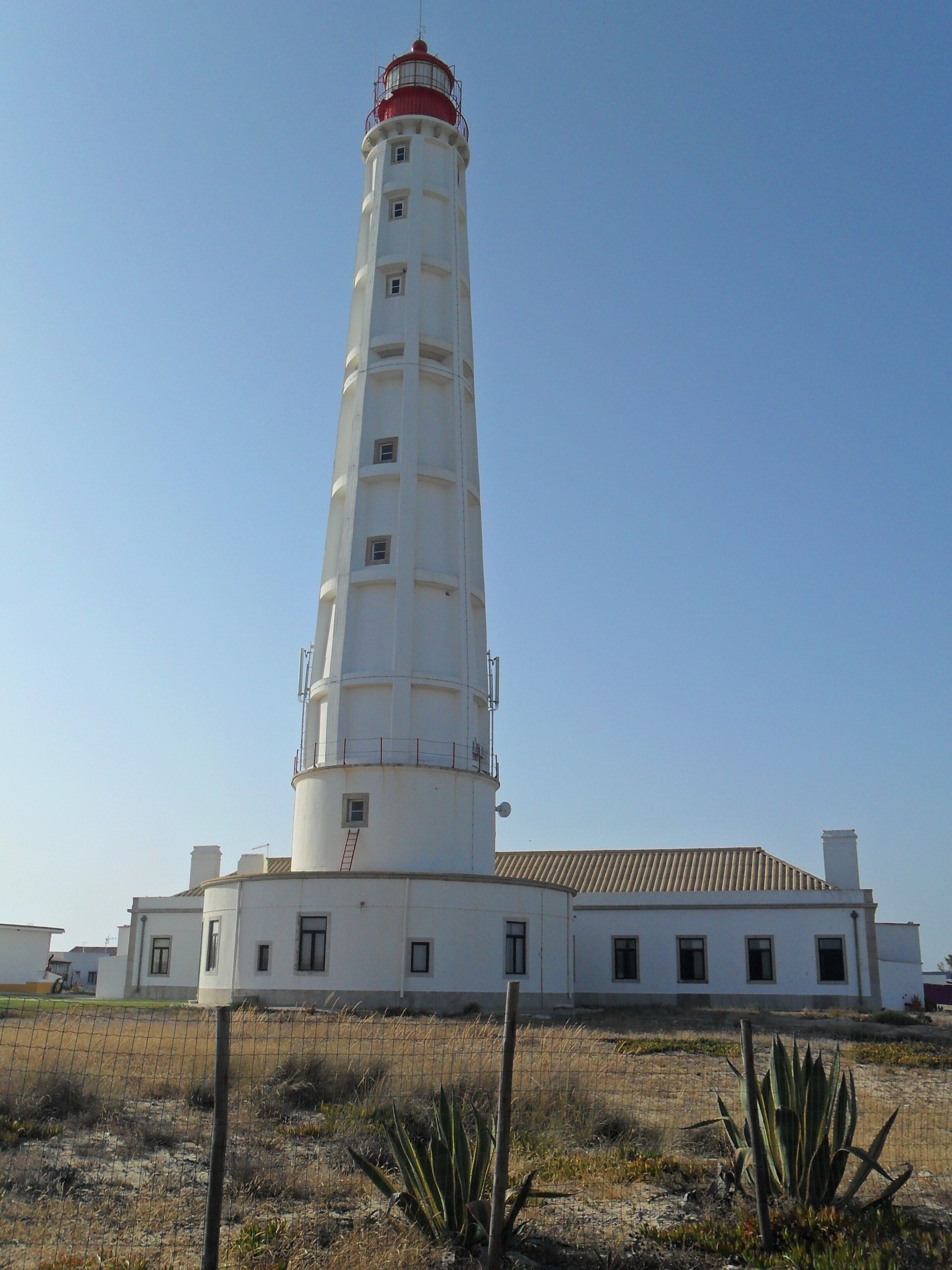 Un grand phare blanc, surmonté d’une partie rouge, entouré d’une zone délimitée par une clôture et où la végétation est clairsemée.