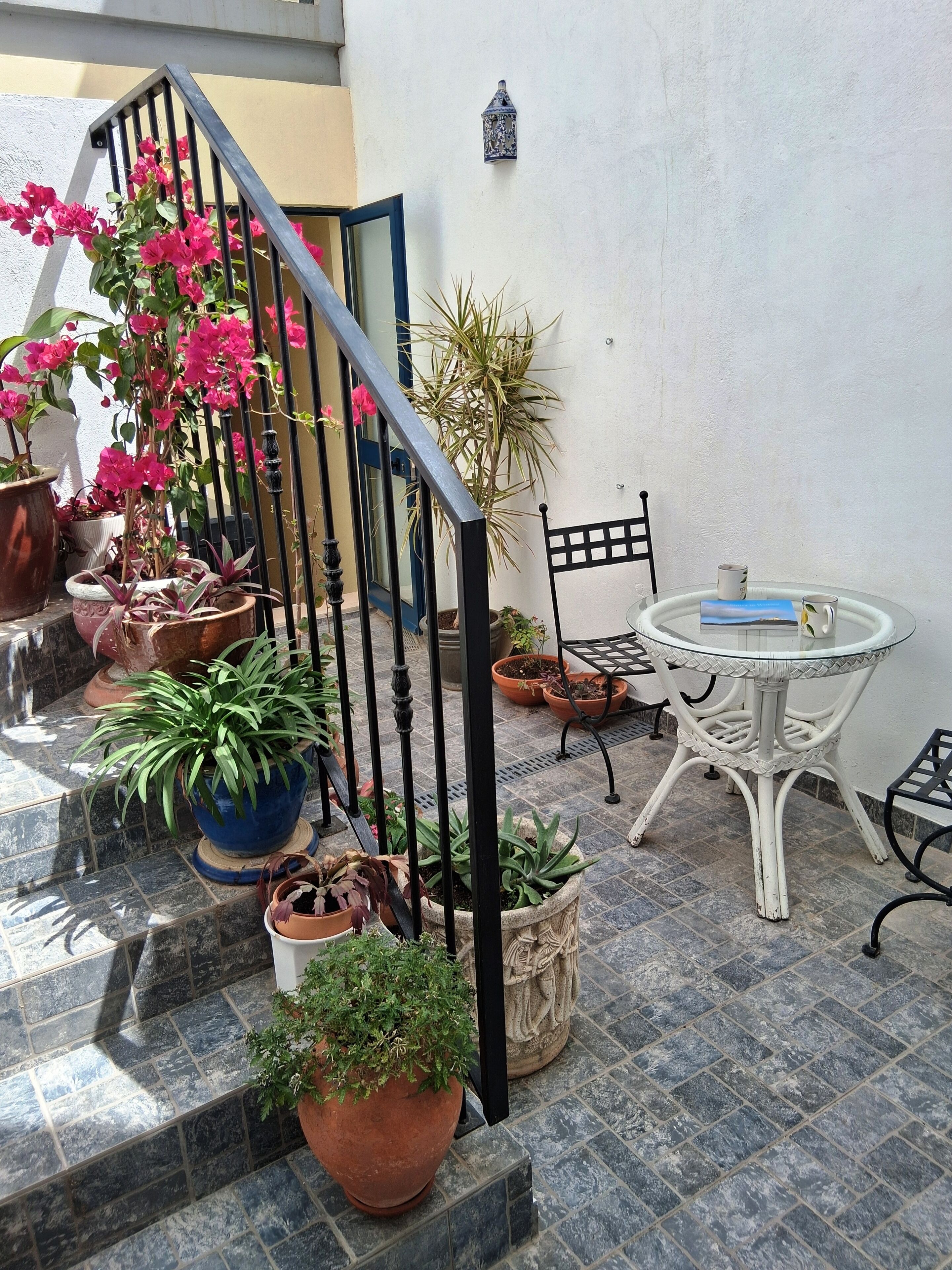 Una terraza con plantas en macetas, una escalera, una mesa redonda con sillas y un farol fijado a la pared.