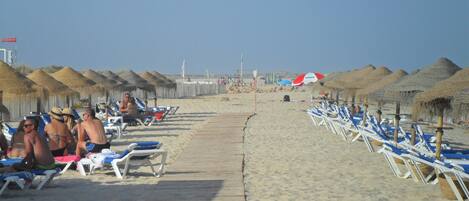 A beach with rows of sun loungers and thatched umbrellas, a wooden walkway, and a clear sky.