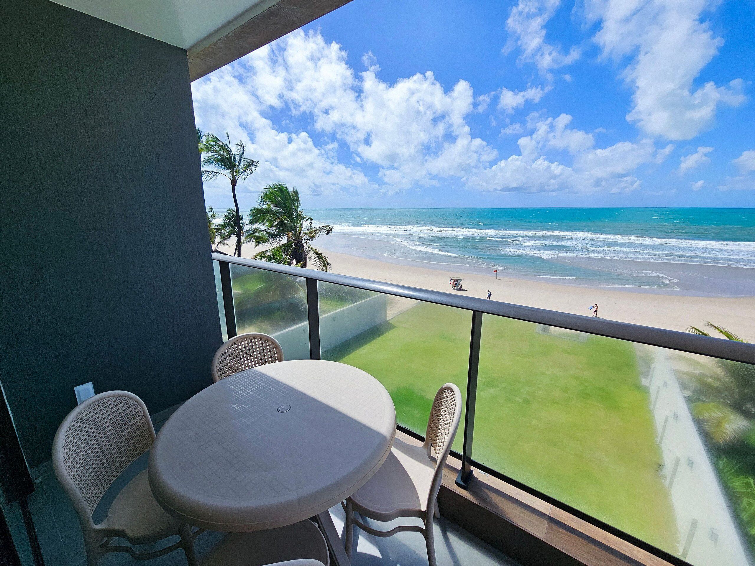 Balcón con mesa y sillas con vista a una playa con palmeras y el océano.