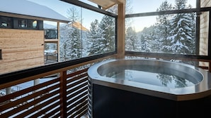 Eine moderne Holzhütte mit einem Außenbereich aus Holz, einer von Glas umschlossenen Badewanne und Blick auf schneebedeckte Bäume.