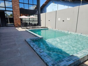 A swimming pool with tiled edges, a patio area with chairs and umbrellas, and a building with large windows. - Atlantic Residence Carneiros (Tamandaré)