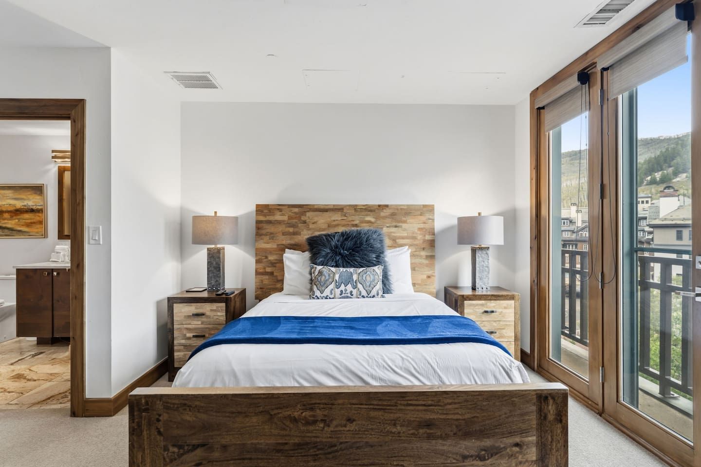 Un dormitorio moderno con cabecera de madera, una cama grande con ropa de cama blanca, dos mesitas de noche con lámparas y vistas a la ciudad a través de amplios ventanales.