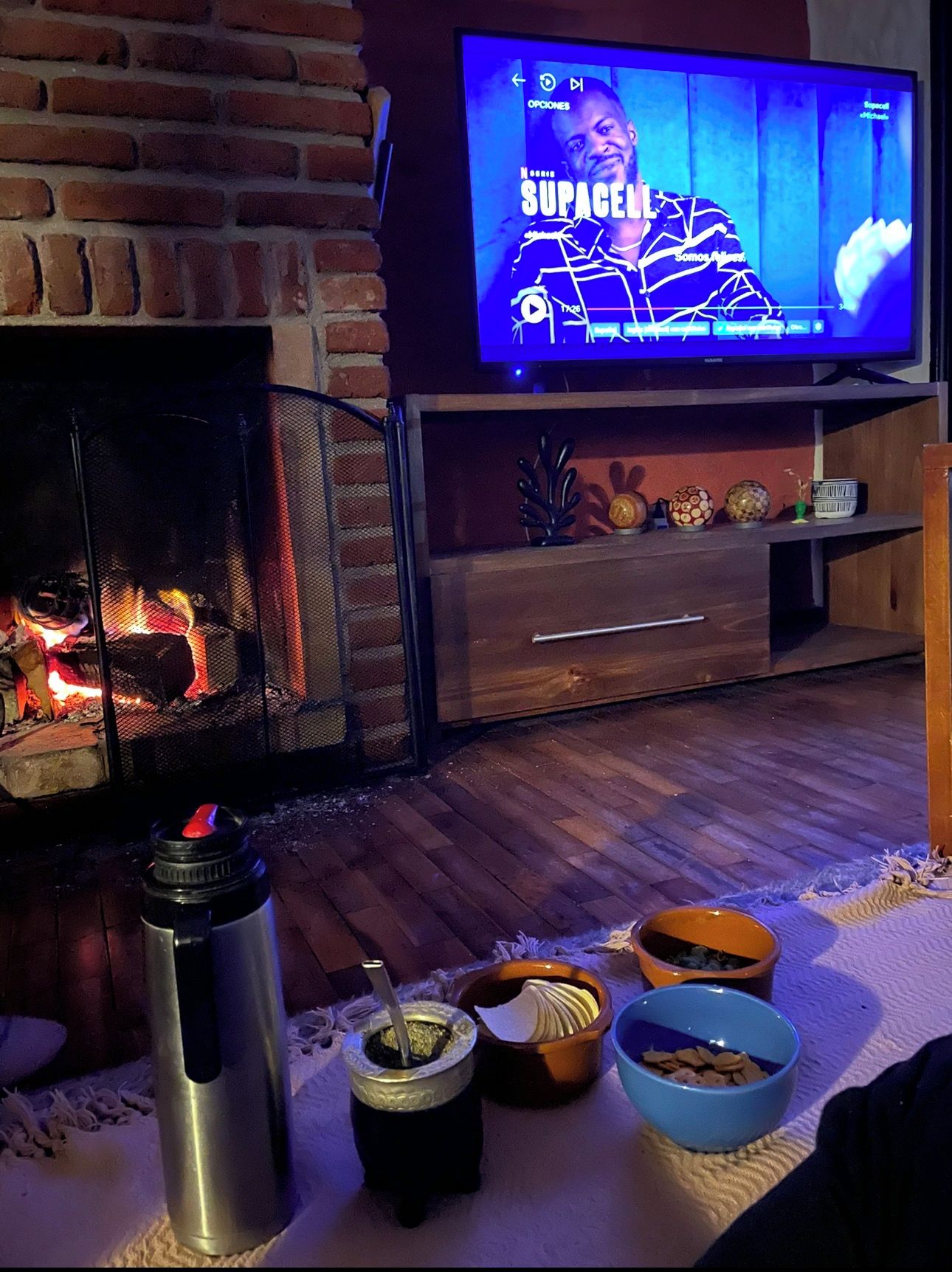 Una sala de estar con una televisión mostrando un programa, una chimenea y una mesa con varios objetos, incluyendo un termo y cuencos.