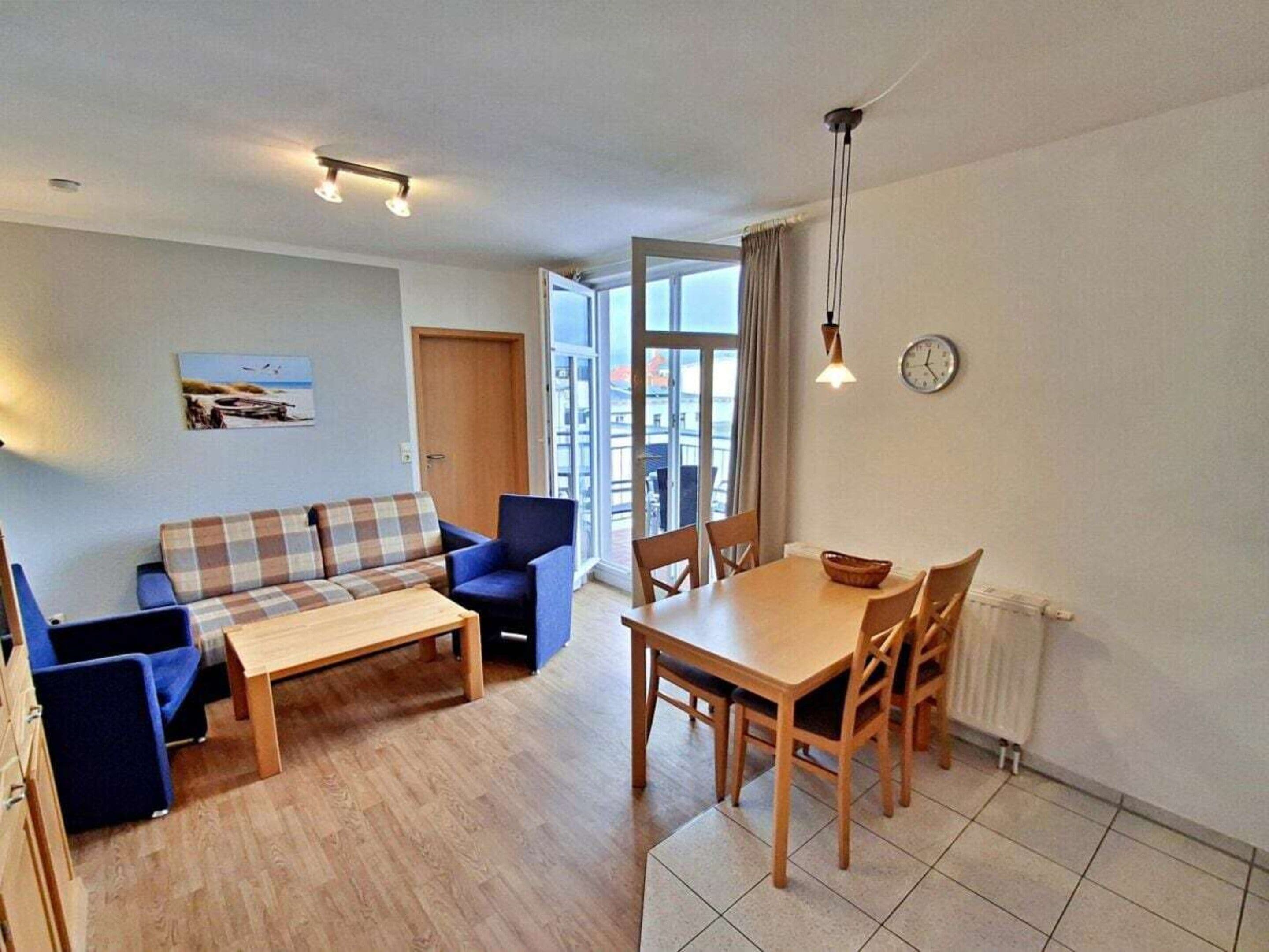 Ein Wohnzimmer mit einer Couch, einem Couchtisch und einem Esstisch mit Stühlen. Es gibt ein Fenster mit Vorhängen, eine Uhr an der Wand und ein Bild an der Wand.