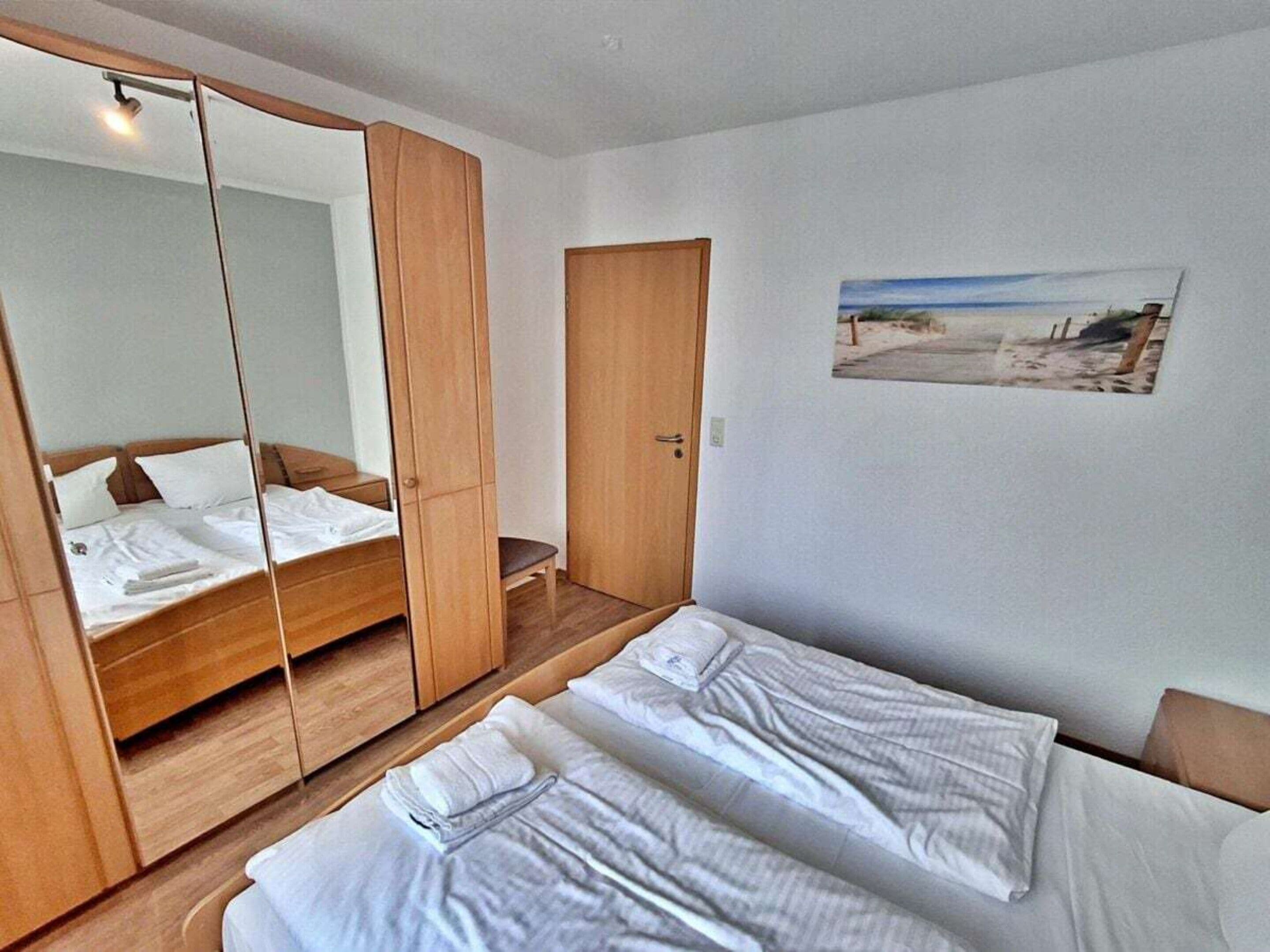 Hotellihuone, jossa on kaksi sänkyä, suuri peili, puinen vaatekaappi ja seinällä oleva taulu, jossa on kuvattuna rantamaisema.