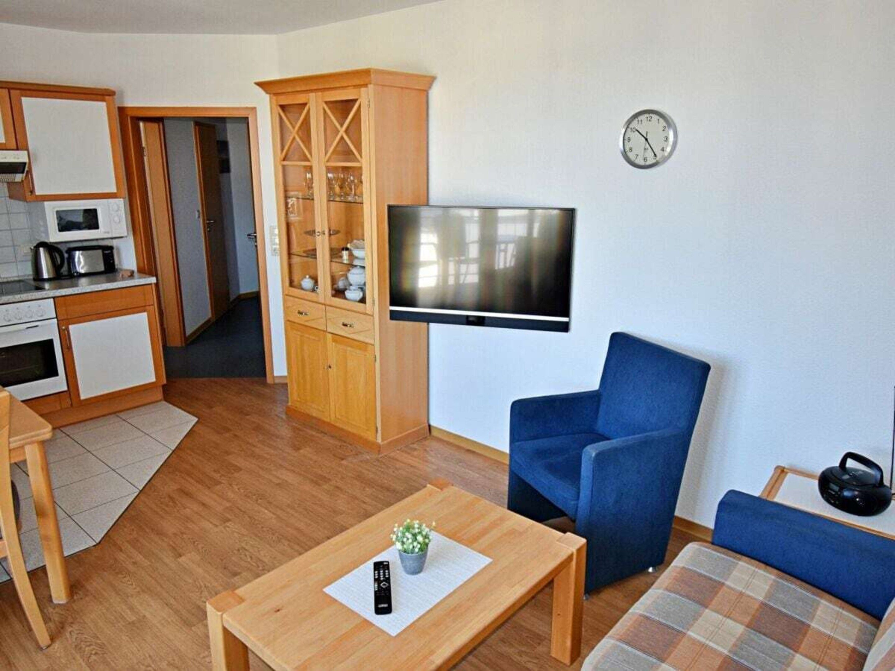 Un espacio habitable compacto con kitchenette, un televisor de pantalla plana montado en la pared, un sillón azul, un sofá a cuadros y una mesita con un control remoto y una planta.