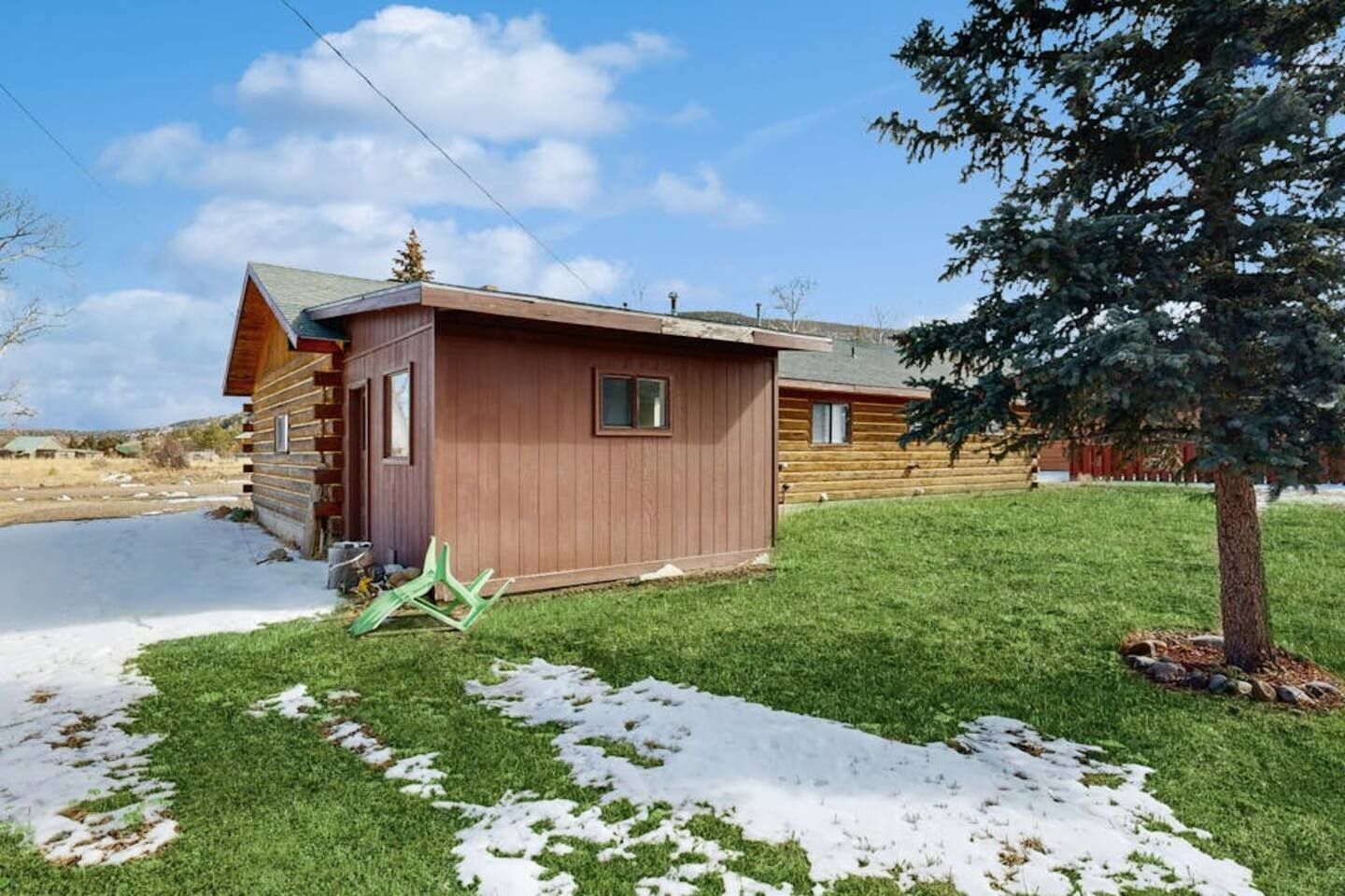 A log cabin with a small attached shed, a grassy yard, and a snow-covered ground.
