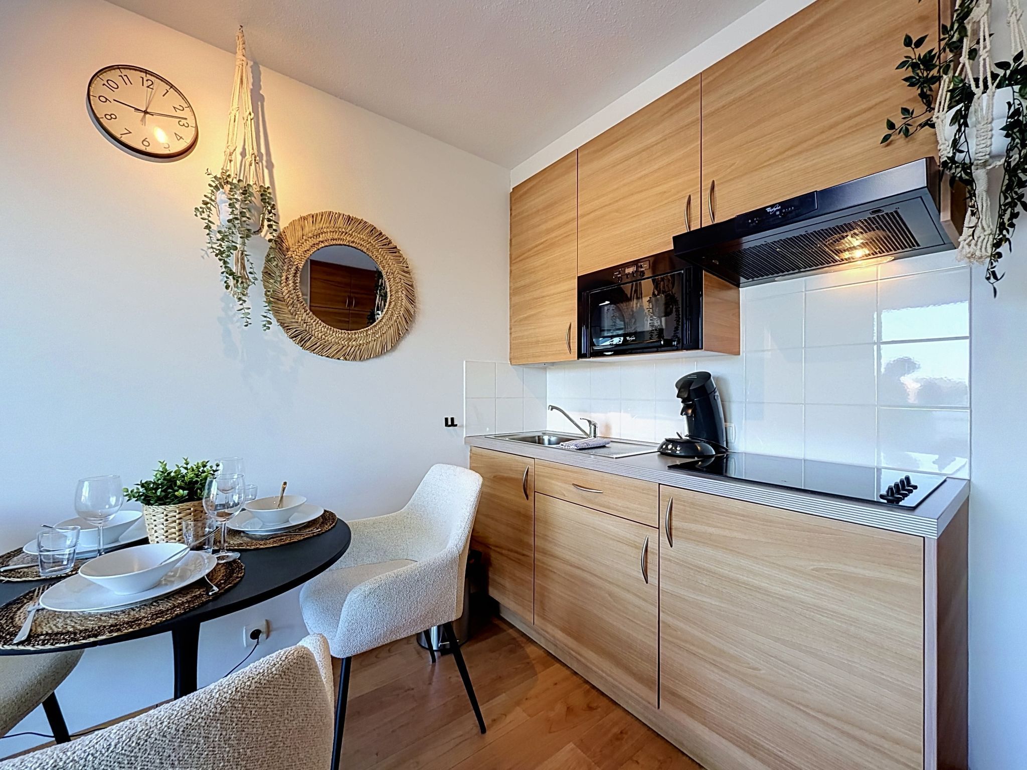 A compact kitchen with wooden cabinets, a black microwave, and a round woven mirror.