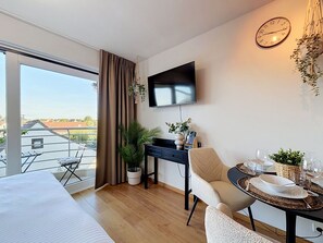 A modern hotel room with a large window, a flat-screen TV, a dining table set for two, and a small desk with a chair. - Sea You @ Blutsyde by Interhome (Bredene)