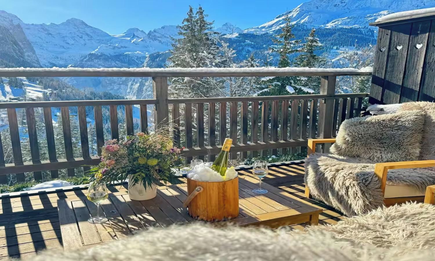 A wooden deck with a view of snow-capped mountains, a bottle of champagne, a glass, and a fluffy chair.