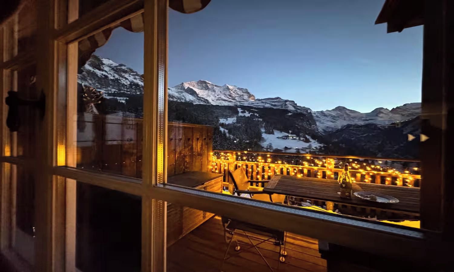 Une vue depuis une fenêtre donnant sur une terrasse avec une balustrade en bois, une table et des chaises, et des montagnes enneigées en arrière-plan.