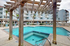 A pool area with a wooden pergola structure, a pool, and a building with balconies in the background. - Seascape Condos - A SeaScape Paradise #1101 (Galveston)