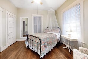 A bedroom with a bed, a bedside table, a chair, a window with curtains, a door, and a ceiling fan. - Castaway Cottage - Big Backyard & Dog-Friendly (Galveston)