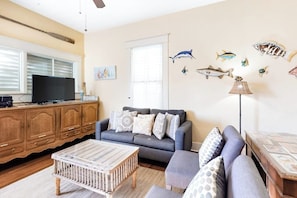 A living room with a gray sofa, a wooden coffee table, and a television on a wooden cabinet. - Castaway Cottage - Big Backyard & Dog-Friendly (Galveston)