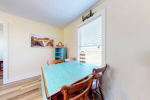 A dining area with a large table and chairs, a window with blinds, a wall-mounted shelf, and a painting. - Beach to Bay Getaway (Galveston)