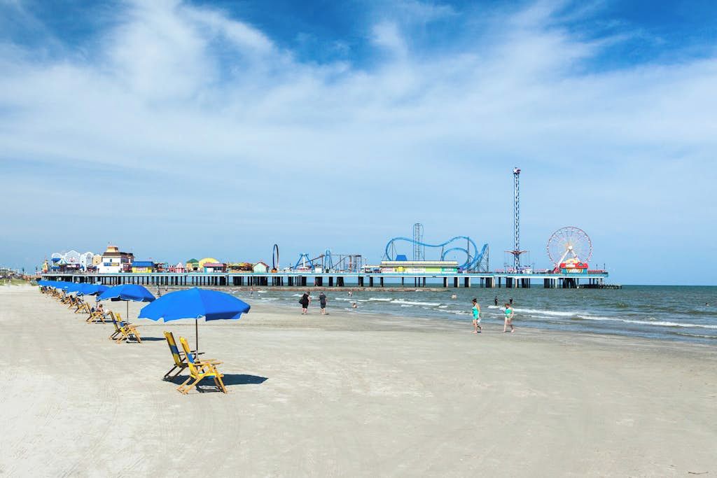 Ein Strand mit Liegestühlen und Sonnenschirmen, ein Steg mit Fahrgeschäften und das Meer im Hintergrund.