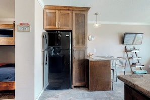Cocina compacta con armarios de madera, un refrigerador negro, una pequeña zona de comedor con sillas y un televisor montado en la pared.