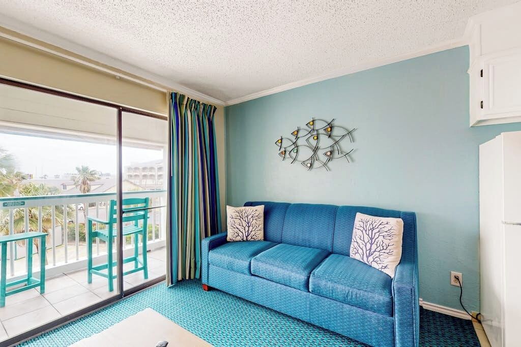 Ein Zimmer mit einem blauen Sofa, einem gemusterten Teppich, einem Balkon mit Stühlen und einem Wanddekor.