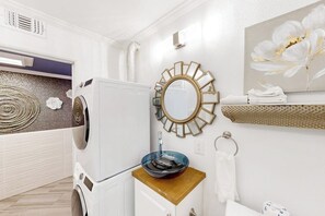 A modern bathroom with a washing machine, a sink with a blue basin, a decorative mirror, and a towel rack. - The Waves: unit A - Beach Duplex, Full Kitchen (South Padre Island)